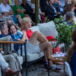 Ludzie siedzą wygodnie na sofie na świeżym powietrzu. Wokół rośliny w donicach, a w tle widoczny tłum. Scena pełna kolorów: czerwień, zieleń i niebieski.