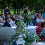 Grupa osób siedzi przy białych stołach na zewnątrz w lesie. Stoliki zdobią zielone rośliny. Ubrania są kolorowe, głównie w odcieniach czerwieni i zieleni.