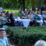 Grupa osób siedzi na spotkaniu plenerowym, otoczona zielenią parku. Na białym stole stoją świece. Tło to drzewa i trawa.