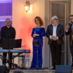 Na zdjęciu grupa elegancko ubranych osób na scenie. W tle fioletowe i czerwone oświetlenie. Obok nich stoi fortepian i mikrofon.