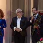 Na zdjęciu są trzy osoby na scenie. Kobieta w niebieskiej sukni, mężczyzna z bukietem róż i starszy mężczyzna w ciemnym garniturze. Tło jest jasne, z czerwonym oświetleniem.