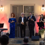 Grupa sześciu osób stoi na scenie, trzy przemawiają do mikrofonów. Kobieta w niebieskiej sukni na tle oświetlenia w czerwieni i fiolecie. Tło to jasna ściana z oknami, przy schodach są kwiaty i instrumenty muzyczne.