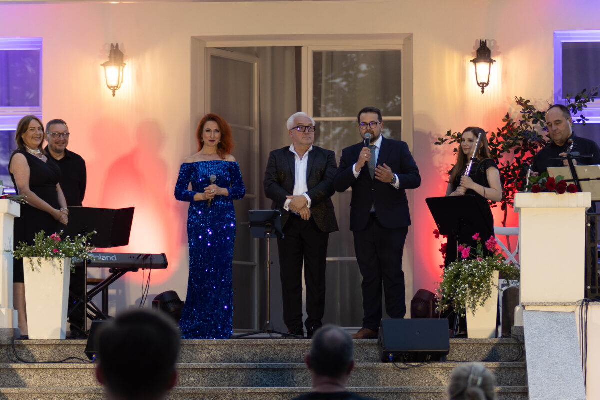 Grupa sześciu osób stoi na scenie, trzy przemawiają do mikrofonów. Kobieta w niebieskiej sukni na tle oświetlenia w czerwieni i fiolecie. Tło to jasna ściana z oknami, przy schodach są kwiaty i instrumenty muzyczne.