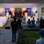 Na zdjęciu jest wieczorna uroczystość przed budynkiem z czerwonym dachem. Grupa ludzi stoi na schodach sceny, podświetlona niebieskim i fioletowym światłem. Widzowie oglądają wydarzenie na kamiennej ścieżce otoczonej zielenią