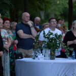 Grupa ludzi stoi na zewnątrz podczas wydarzenia. Na pierwszym planie stół z zielonymi roślinami i białym obrusem. W tle zielone drzewa.