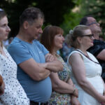 Zdjęcie przedstawia pięć osób stojących w rzędzie na tle zielonych drzew. Osoby patrzą w jedną stronę, a jedna z nich trzyma telefon. Stroje są w jasnych kolorach z elementami wzorów.