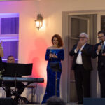 Na zdjęciu jest grupa osób elegancko ubranych na scenie. Kobieta w niebieskiej sukni trzyma mikrofon. Tło to jasna ściana i okno z fioletowym podświetleniem.