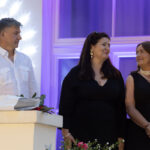 Trzy osoby stoją na tle białej i fioletowej ściany. Dwie kobiety w czarnych strojach, mężczyzna w białej koszuli. Obok bukiet kwiatów.