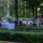 Ludzie siedzą w ogrodzie, oglądają wydarzenie. Naturalne oświetlenie, zielone tło drzew. Stół z białym obrusem i roślinami.