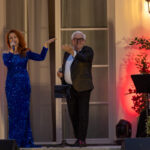 Na scenie kobieta w niebieskiej sukni i mężczyzna w marynarce. W tle czerwone oświetlenie i rośliny. Obok siedzi osoba przy pulpicie.