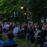 Otwarta przestrzeń, ludzie siedzą na krzesłach w parku. Zieleń i latarnie w tle tworzą wieczorną atmosferę. Widać stoły z dekoracjami.