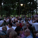 Wieczorne spotkanie na świeżym powietrzu z ludźmi przy białych stołach, w otoczeniu drzew i lamp.