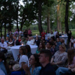 Grupa ludzi siedzi na krzesłach w parku podczas wieczornej imprezy z lampionami. Otaczają ich drzewa, a tło jest zielone. Stoły pokryte białymi obrusami, na stole świeczki.