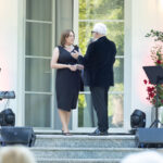 Para rozmawia na schodach w eleganckich strojach. Tło to jasna fasada z oknem i lampami. Obok stoją rośliny i instrumenty muzyczne.