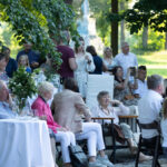 Grupa ludzi siedzi przy białych stołach na świeżym powietrzu w parku, otoczona zielenią. Ubrani są kolorowo, a atmosfera jest swobodna. W tle widoczny jest zalesiony krajobraz.