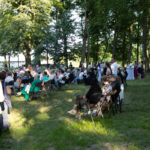 Na zdjęciu widać ludzi siedzących na krzesłach w parku. W tle widać drzewa i stoliki koktajlowe z białymi obrusami. Słońce rzuca cienie, a ludzie są w jasnych ubraniach.