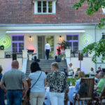 Na zdjęciu widać osób w ogrodzie przed białym dworkiem z czerwonym dachem. Na schodach stoi mężczyzna w bieli. W tle świecą kolorowe reflektory.