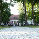 Ścieżka z białego żwiru prowadzi przez park do dużego, białego domu z czerwonym dachem. W tle zgromadzeni ludzie. Dookoła zielone drzewa.