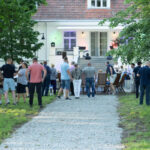 Spotkanie na świeżym powietrzu przy białym budynku z czerwonym dachem. Ludzie stoją na żwirowej ścieżce i rozmawiają. W tle drzewa i zielona trawa.