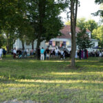 Grupa ludzi zgromadzona przed białym budynkiem z czerwonym dachem. Drzewa i trawa na pierwszym planie. Impreza na zewnątrz.