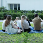 Grupa ludzi siedzi na trawie na kocu, w tle białe budynki. Jedna osoba ma jasną sukienkę w kwiaty. Wokół dużo zieleni.