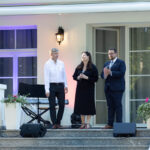 Trzy osoby stoją na schodach przed białym budynkiem. Tło tworzą fioletowe światła, okna i drzwi. Obok nich są instrumenty muzyczne i kwiaty.