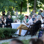 Grupa ludzi siedzi na zewnątrz, przy okrągłych stolikach, otoczona zielenią drzew. Uczestnicy są elegancko ubrani i biją brawo. W centrum znajduje się biały stolik z wazonem kwiatów.