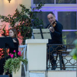 Muzyk gra na skrzypcach, a obok kobieta z fletem na tle zieleni. Żywe kolory z kwiatami na białym parapecie. Ciepłe oświetlenie.