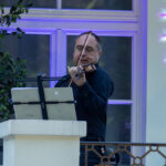 Mężczyzna w ciemnym stroju gra na skrzypcach przed oknem. Tło w białych i niebieskich odcieniach. Na ścianie lśnią wzory.