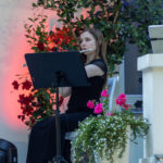 Na zdjęciu kobieta gra na flecie. Siedzi przy nutach na krześle w ogrodzie. W tle kwiaty w donicach, czerwone i różowe.