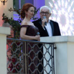 Na zdjęciu elegancko ubrany duet z mikrofonami na tle jasnej fasady budynku. Ona w czerwonej sukni, on w czarnym garniturze. W tle rośliny oraz dekoracyjne światło.