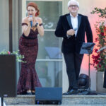 Na zdjęciu są dwie osoby śpiewające na scenie. Kobieta ma czerwoną sukienkę, mężczyzna czarny garnitur. Tło stanowi jasna ściana i okno.