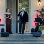 Na zdjęciu jest koncert na świeżym powietrzu. Kobieta w ciemnej, błyszczącej sukni i mężczyzna w czarnym garniturze śpiewają. Tło to jasna ściana i zieleń, z czerwonymi światłami.