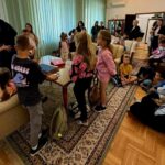 Grupa dzieci w różnorodnych kolorowych ubraniach siedzi i stoi w salonie z zielonymi zasłonami oraz drewnianą podłogą. Dzieci wokół stołu, na kanapach i dywanie, otoczone jasnymi ścianami. W tle dorośli obser