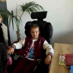 Dwie dziewczynki w biurze, jedna siedzi na czarnym fotelu. Obok stoi z palmą w tle. Drewniane biurko z teczką po prawej.