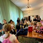 Grupa dzieci i dorosłych w pokoju z zielonymi zasłonami, drewnianym stołem i żyrandolem. Dzieci słuchają kobiety. Atmosfera edukacyjna.