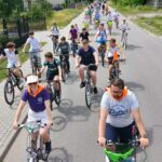 20250629_1343021 Grupa rowerzystów jedzie ulicą w słoneczny dzień. Widoczne zielone krzewy i domy w tle. Różnorodne stroje, kaski i plecaki.