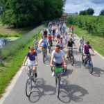 20250629_141506 Grupa ludzi jedzie rowerami po asfaltowej drodze. Po obu stronach drogi znajdują się zielone drzewa i trawa. Tło jest jasne z niebieskim niebem i chmurami.