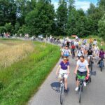 20250629_141540 Grupa rowerzystów na wiejskiej drodze otoczonej zielenią, słońce świeci jasno, w tle las i niebo.