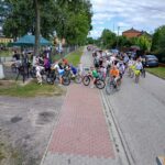 20250629_143444 Grupa rowerzystów na ścieżce, otoczona zielenią i domami, pod niebieskim, zachmurzonym niebem. Alpinizm, czerwony bruk na drodze.