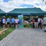 20250629_143638 Ludzie stoją w kolejce pod zielonym namiotem na targu. W tle widoczne są chmury i trawa. Obok wisi baner Gmina Dobre.