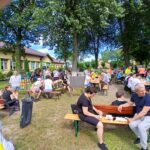 20250629_145111 Na zdjęciu widać ludzi siedzących na ławkach pod drzewami na pikniku. Tło stanowi budynek z kremową elewacją. Dominuje zielona i niebieska kolorystyka.