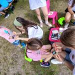 20250629_145215 Grupa dzieci i dorosłych na trawie maluje farbami. Ubrania mają jasne kolory, a w tle widać stół z osobami. Atmosfera plenerowa.