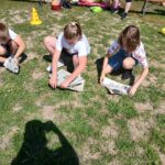 20250629_150701 Trójka dzieci w jasnych koszulkach czyta gazety na trawie. Za nimi widać ludzi i kolorowe pachołki. Słoneczny dzień.