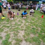 20250629_152143 Na trawie klęczą trzy osoby, skupione na zadaniu, wokół ludzi. Na drugim planie znajduje się zielony namiot i stoły. Tło jest zielone i żywe, z czerwonym pachołkiem po prawej.