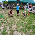 20250629_152247 Grupa ludzi na trawie, biorących udział w aktywności. Zielony namiot w tle, kolorowe stożki na ziemi. Dzień słoneczny, atmosfera piknikowa.