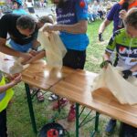 20250629_153821 Na zdjęciu kilka osób szyje przy drewnianych stołach na świeżym powietrzu. Ubrani są kolorowo, wokół widać rowery. W tle grupka ludzi obserwuje wydarzenie.