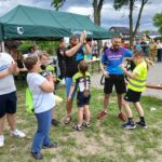20250629_154000 Grupa ludzi stoi na trawie, obok zielonego namiotu. Kilka osób w sportowych ubraniach, w tle są drzewa i budynki. Atmosfera przypomina festyn lub wydarzenie sportowe.
