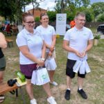20250629_154008 Trzy osoby w białych koszulkach stoją na trawie, trzymając torby. W tle drzewa i banner. Jasny, pogodny dzień.
