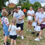 20250629_154041 Grupa ludzi na pikniku, dzieci otrzymują torby z upominkami. Trawa i drzewa w tle, niebieskie i białe ubrania dominują.